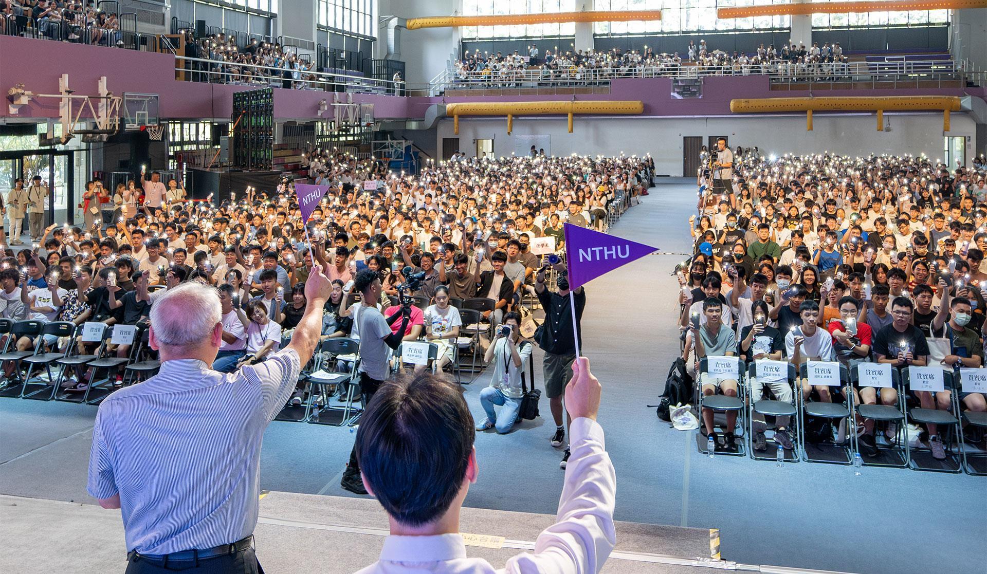 A group of faculty holding up NTHU pennants while the freshmen respond by lighting up their mobile phone flashlights.